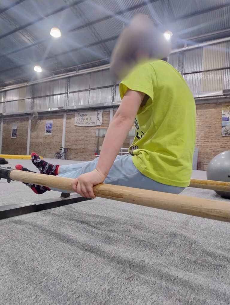 Niño realizando un ejercicio de L-sit en barras paralelas dentro de un gimnasio cubierto.