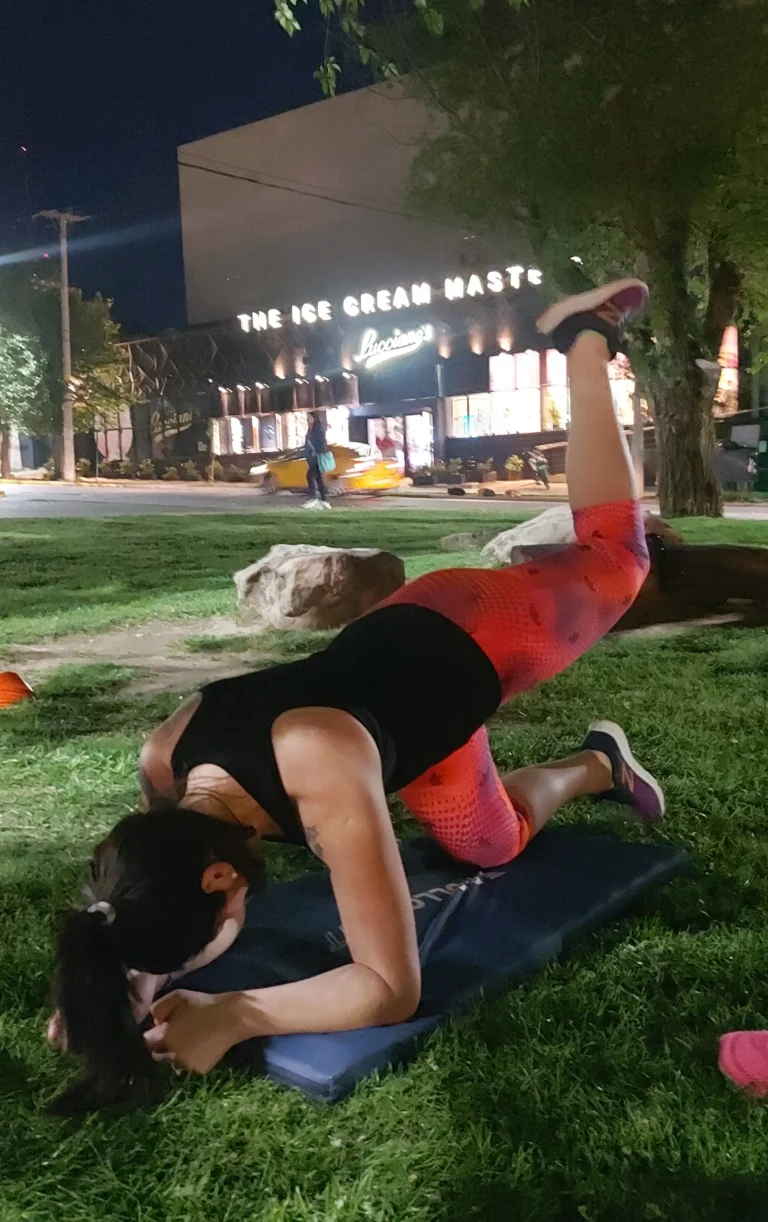Mujer realizando elevación de pierna tipo glute kickback en posición de plancha apoyada en antebrazos, durante un entrenamiento nocturno en un parque.